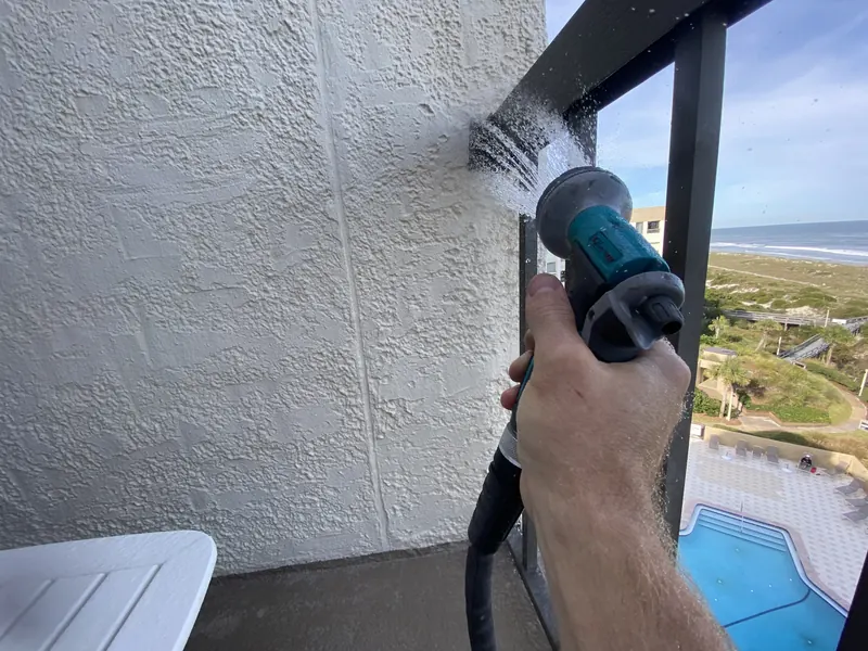 Hand-held nozzle spraying water at the joint where a balcony railing meets a stucco wall, with the ocean in the background