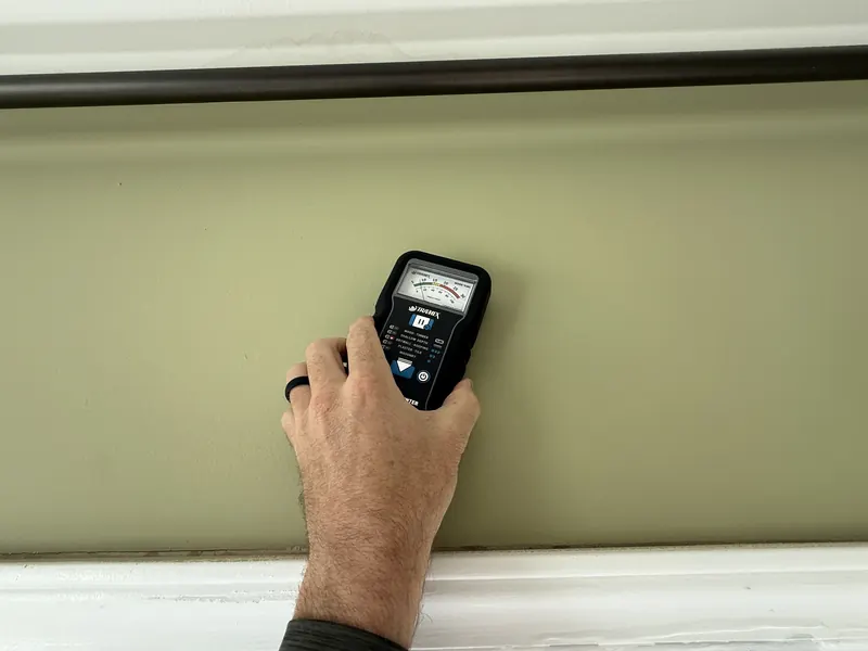 Hand holding a moisture meter against an interior wall below a window